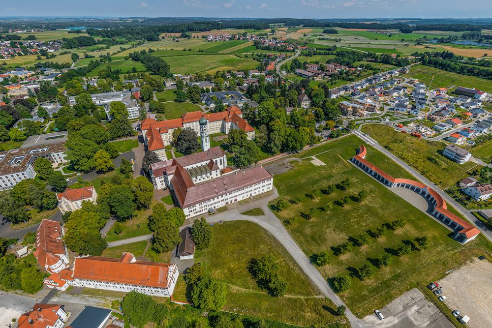 Drohnenaufnahme von Bad Schussenried mit der großen Klosteranlage und Kirche, roten Ziegeldächern, weitläufigem Gelände sowie umliegenden Wohngebieten und Feldern.